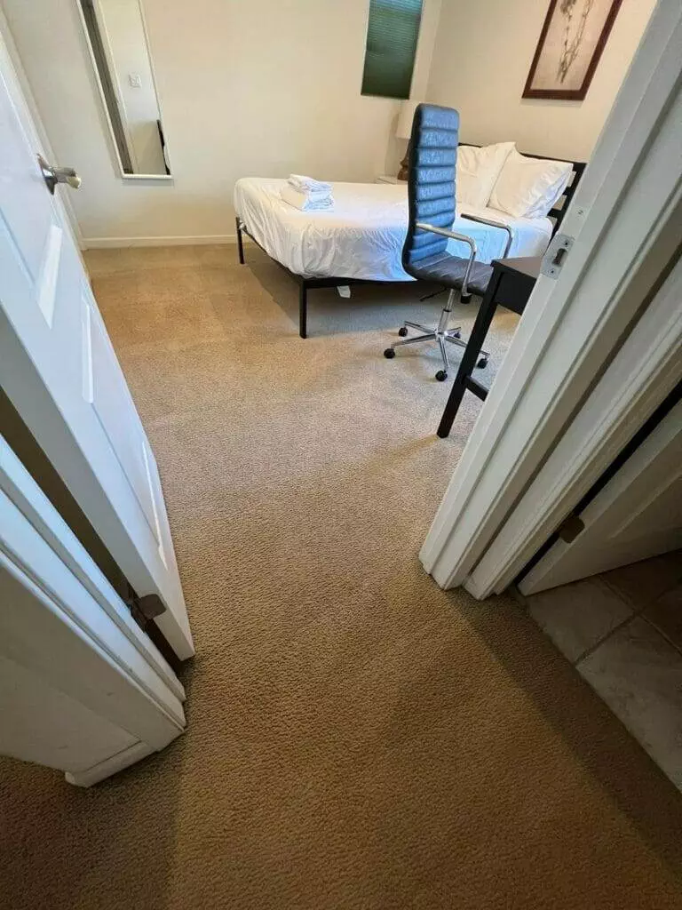 A bedroom with a bed, desk and chair, featuring tough pasta stains on the carpet and a glass of wine on the desk.