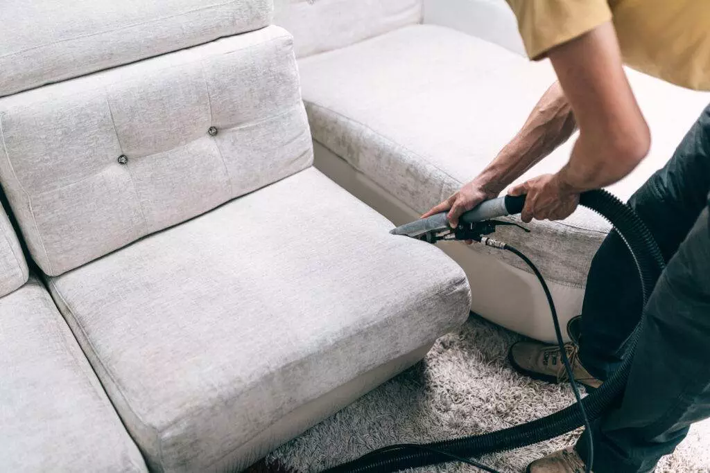 A man expertly cleaning a couch in his living space with a vacuum.
