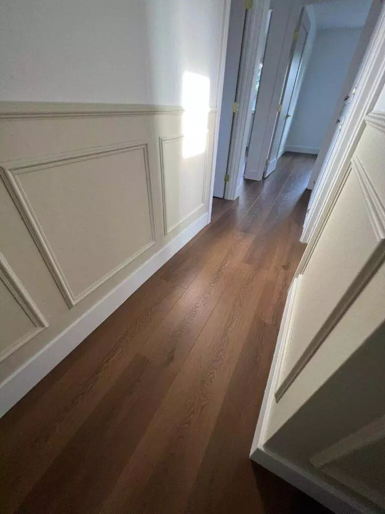 Sunlight streaming through a doorway onto wooden flooring in a hallway with white wainscoting.