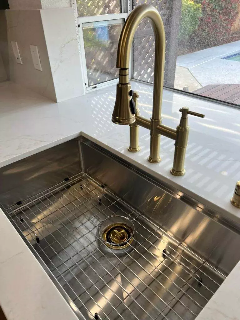 A modern kitchen sink with a stainless steel basin, gold-toned faucet, and a matching drain, set against white countertops.