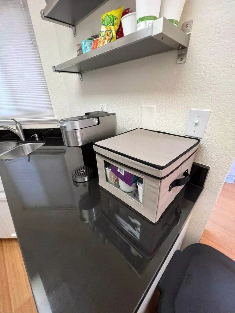A kitchen counter with a fabric storage box, an immaculate coffee maker, floating shelves with items, and a sink visible.