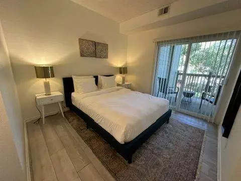 A modern master bedroom with a white double bed, two table lamps, a framed artwork above the bed, and a balcony accessed through a sliding door.