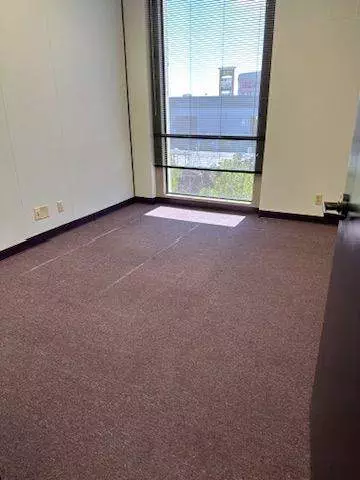 An empty office room with restored purple carpet, a large window with a mesh roller shade, and white walls, showing a distant building outside.