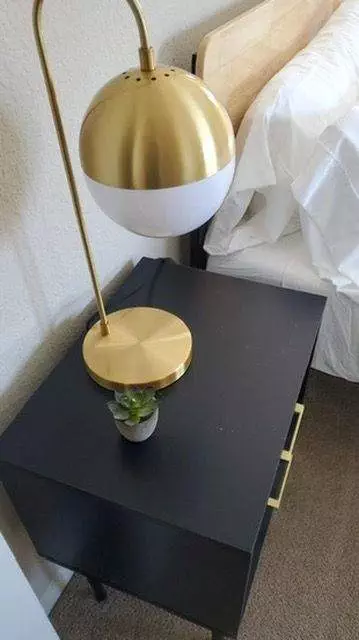 Gold and white desk lamp on a black nightstand with a small potted plant, beside a bed with white bedding in Cupertino.