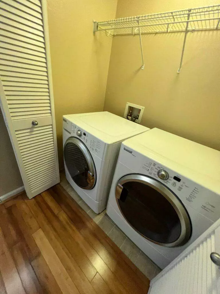 A laundry room with a white Master Clean Service washing machine and dryer set on wooden flooring, and an open louvered door on the left.