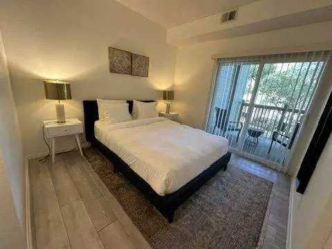 A well-lit modern bedroom with immaculate cleanliness, featuring a large bed, white bedding, side tables with lamps, a framed artwork above the bed, and a balcony with a railing visible through a