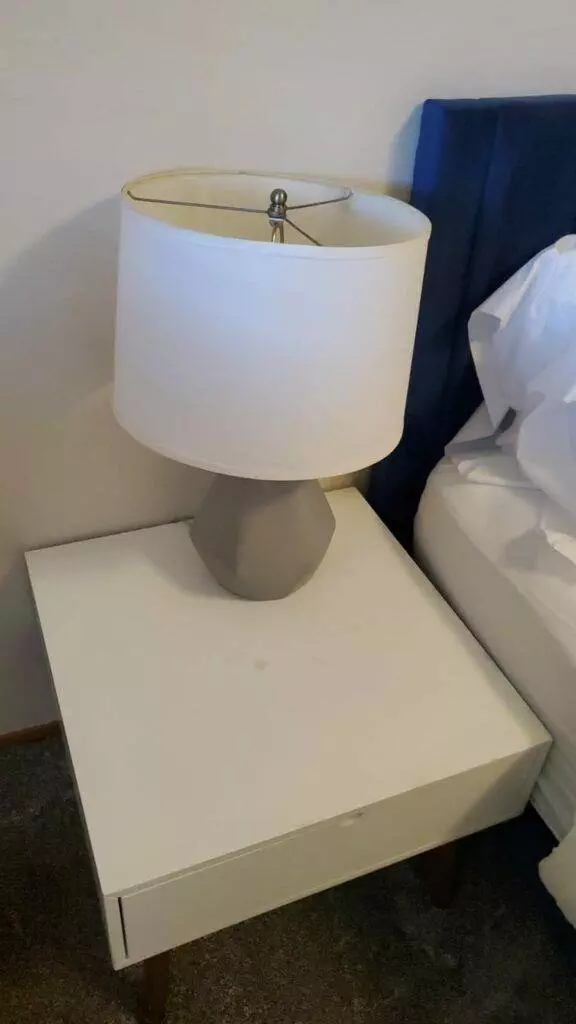 A white table lamp with a cylindrical shade on a white bedside table, provided by Master Clean Service, next to a blue headboard.