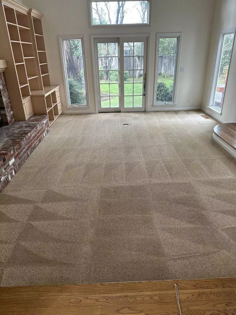 Empty living room with beige carpet by Santa Clara Carpet Cleaning, a red brick fireplace on the left, large windows, and French doors leading to a yard.