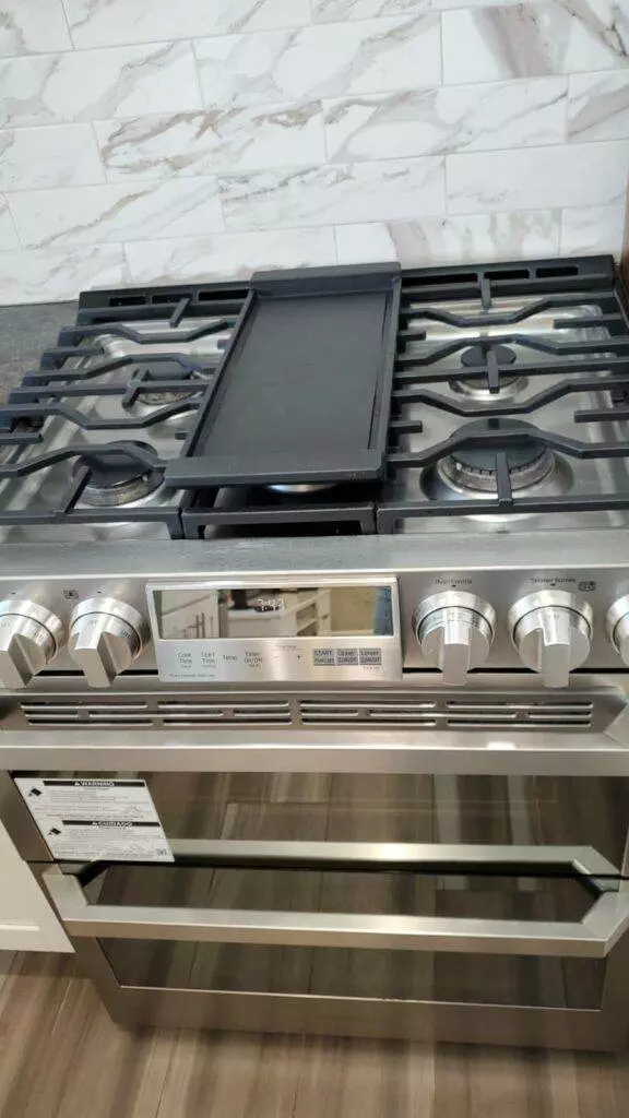 A modern stainless steel gas stove with a central griddle, control knobs, and a digital display, set against a spotless marble backsplash.