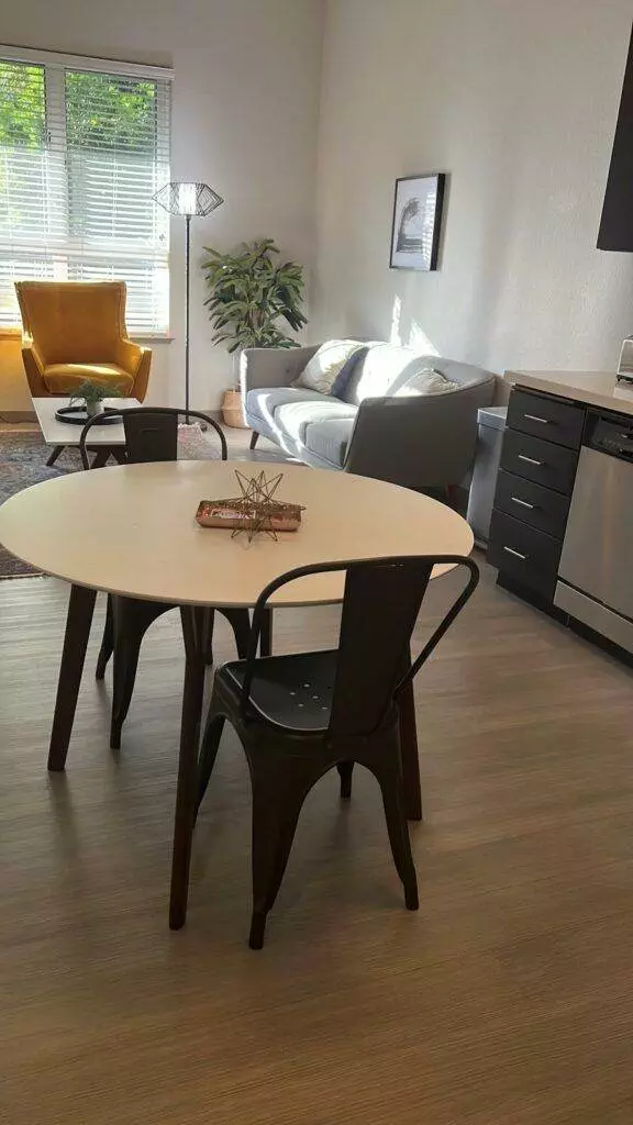 A modern living room with a round white table, three chairs, a sofa, and an adjacent kitchen area visible. Natural light streams through the window. This space has recently benefited from deep cleaning by Master