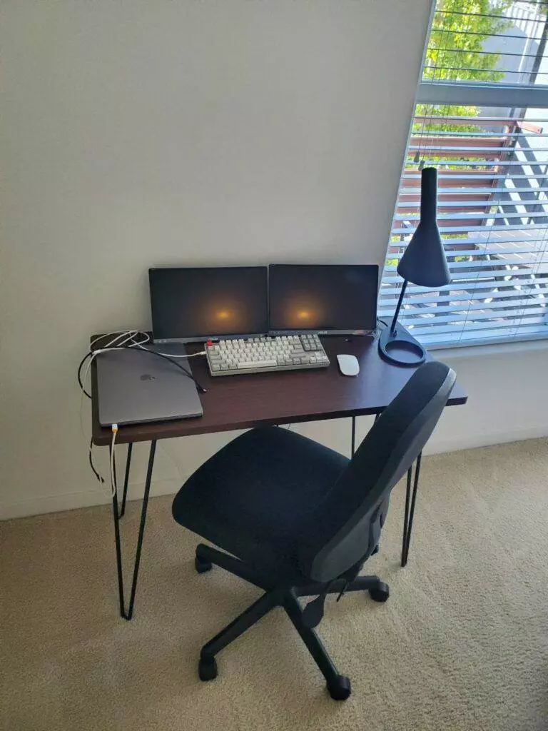 A home office setup featuring a desk with two monitors, a laptop, a mechanical keyboard, and a desk lamp provided by Master Clean Service, next to a window with blinds and a black office chair.