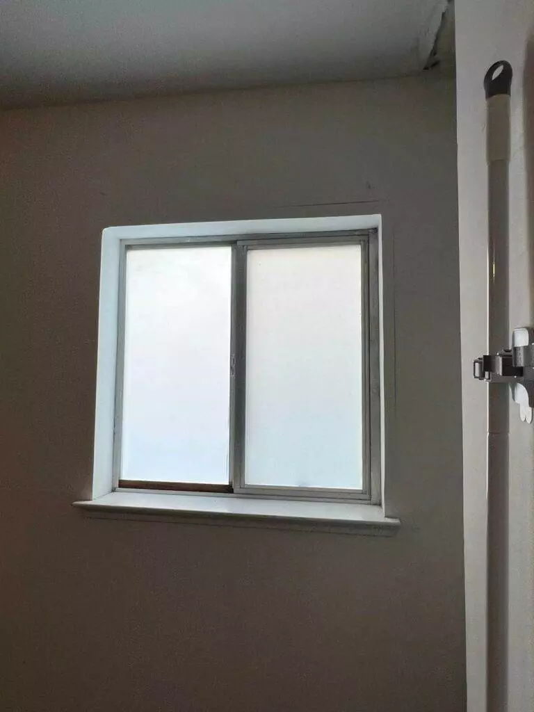 A small, square window with white frames set in a grey wall, partially obscured by a blurry reflection from recent renovation cleaning, next to a black vertical grab bar.