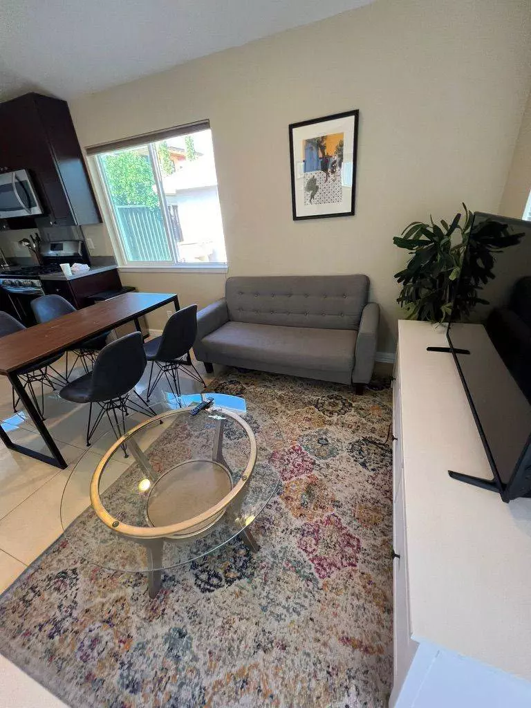 A cozy living room featuring a gray sofa, a wooden dining table with chairs, a glass coffee table, and a colorful area rug, professionally cleaned by Union City Corporate House Cleaning, with a kitchen visible
