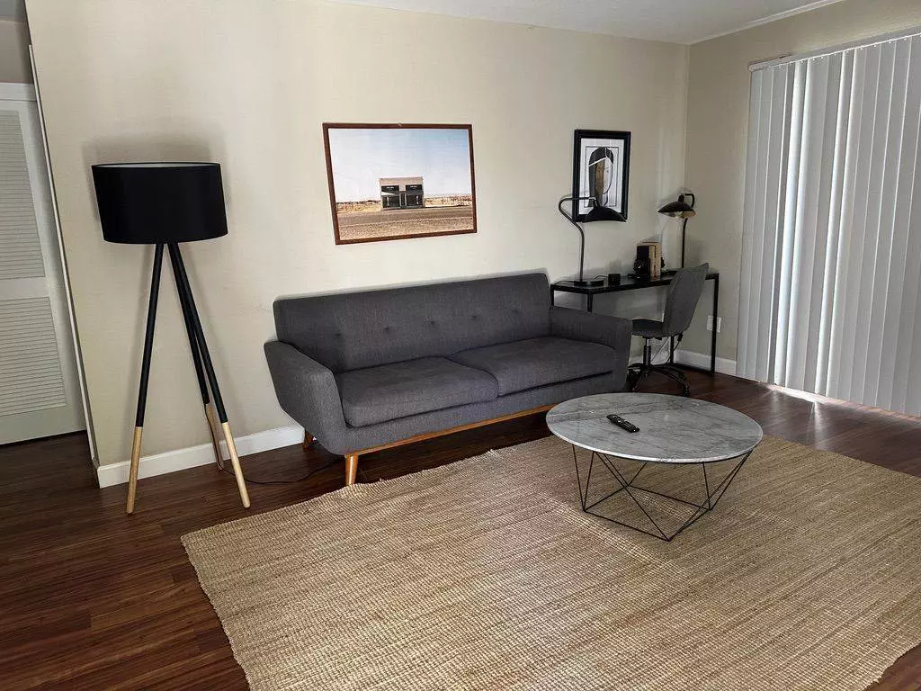 Modern living room in a Cupertino AirBNB with a gray couch, white circular coffee table, and a framed photograph above, hardwood floors, and a small desk in the corner.