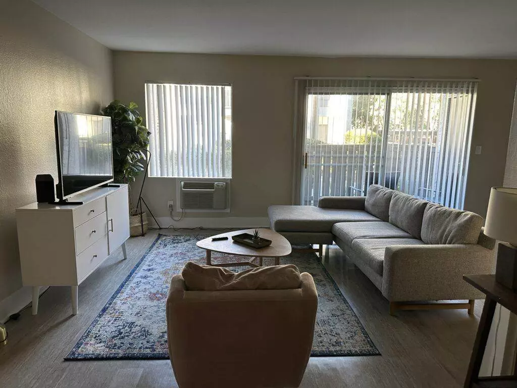 A modern living room featuring a gray sectional sofa professionally treated by Santa Clara Upholstery Cleaning, a television on a white stand, a round wooden coffee table, and a large window with vertical blinds.