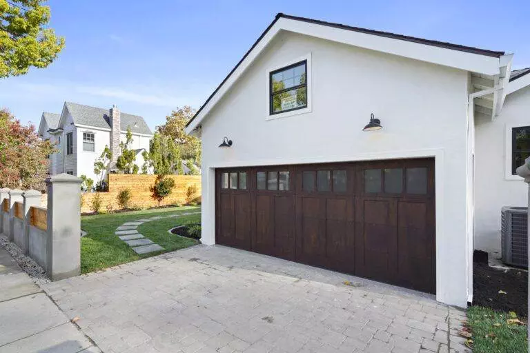 A modern white garage with a dark wooden door, adjacent to a landscaped yard with a paved pathway and a residential house in the background, featured in our post-construction portfolio.