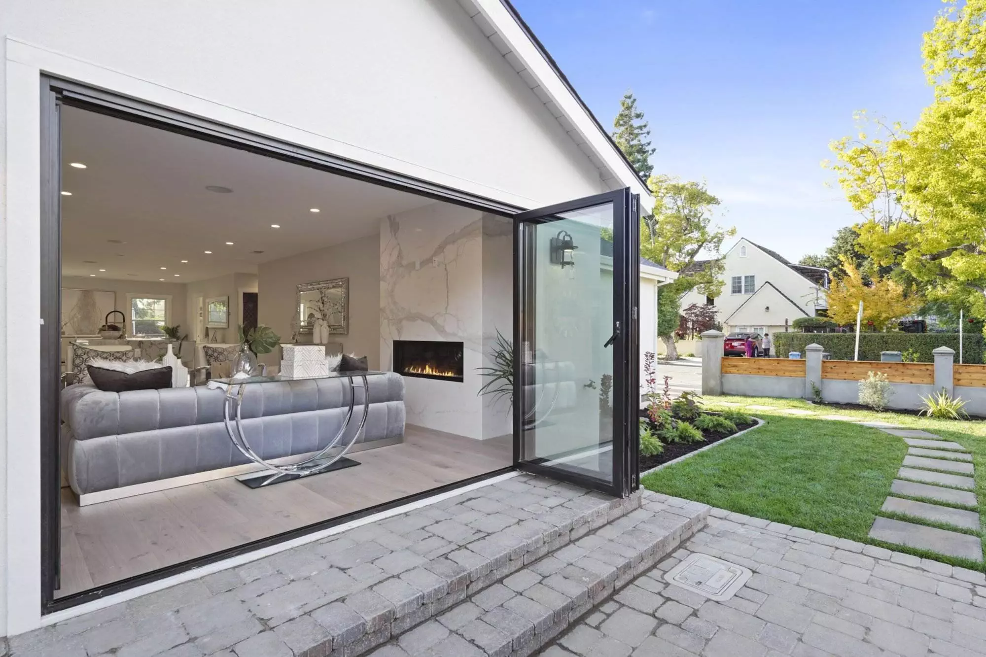 Modern living room visible through large open sliding glass door, featured in a post-construction portfolio, with gray sofas, marble fireplace, and overlooking a landscaped garden.