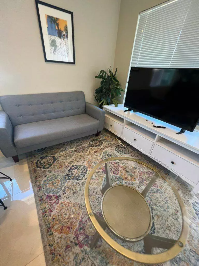 A modern living room featuring a gray upholstered sofa, white TV stand, a colorful rug, a round glass coffee table, a plant, and framed wall art.
