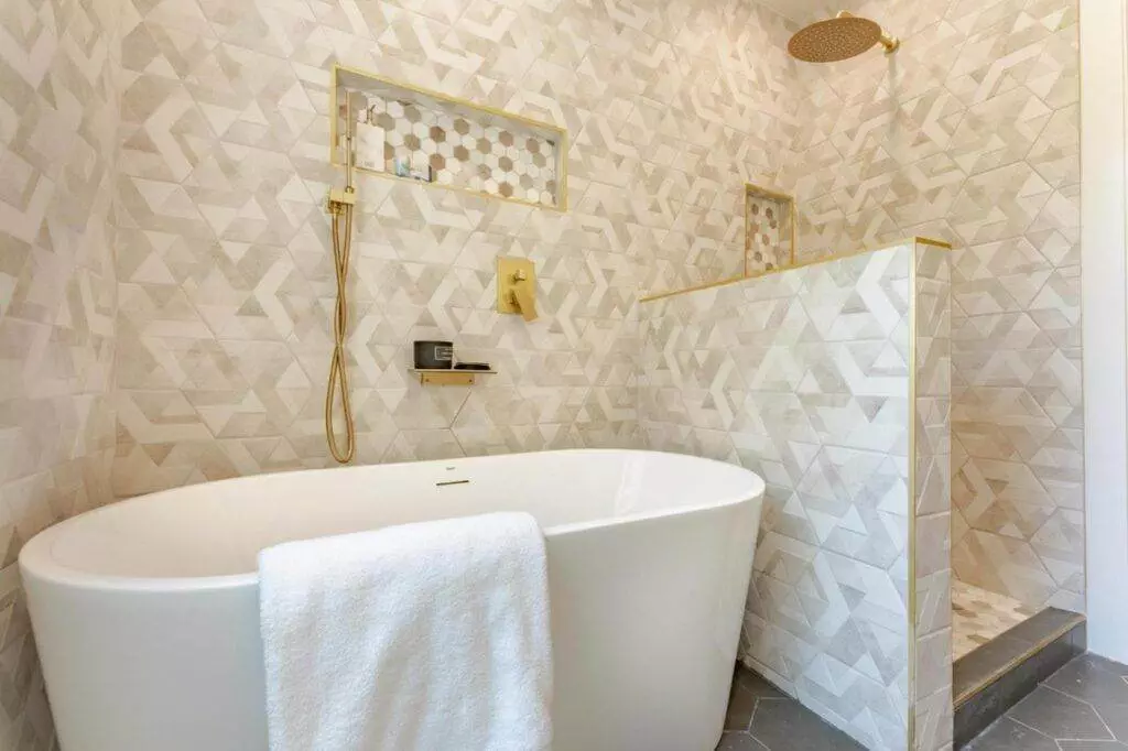 Modern bathroom with a white freestanding tub, herringbone tile walls, and gold fixtures, including a rain shower head from our post-construction portfolio.