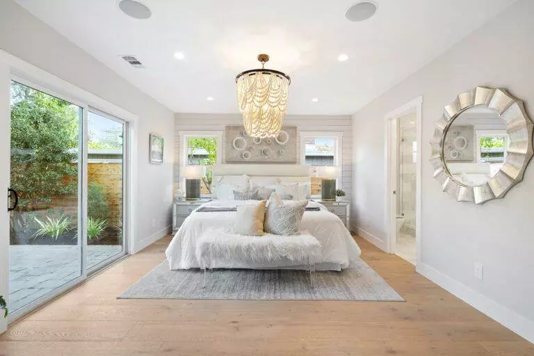 Bright, modern bedroom with a large bed, chandelier, glass doors leading to a garden, and decorative mirrors on the walls newly renovated in our post-construction portfolio.