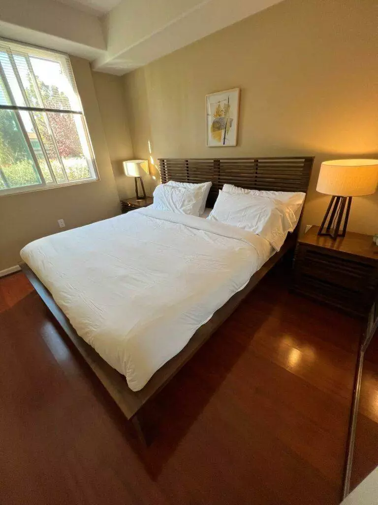 A neatly made bed with white bedding in a bedroom with wooden furniture, a floor lamp, and a window showing greenery outside, maintained by Master Clean Service.