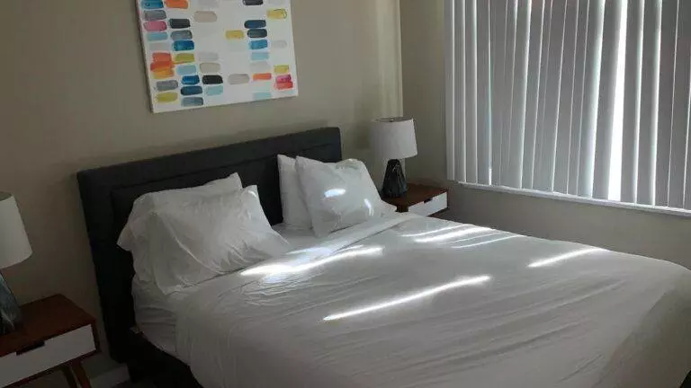 A neatly made bed with white linens in a room with a grey headboard, beside table with a lamp, and a colorful abstract painting above the bed, perfectly prepared for short-term rentals.
