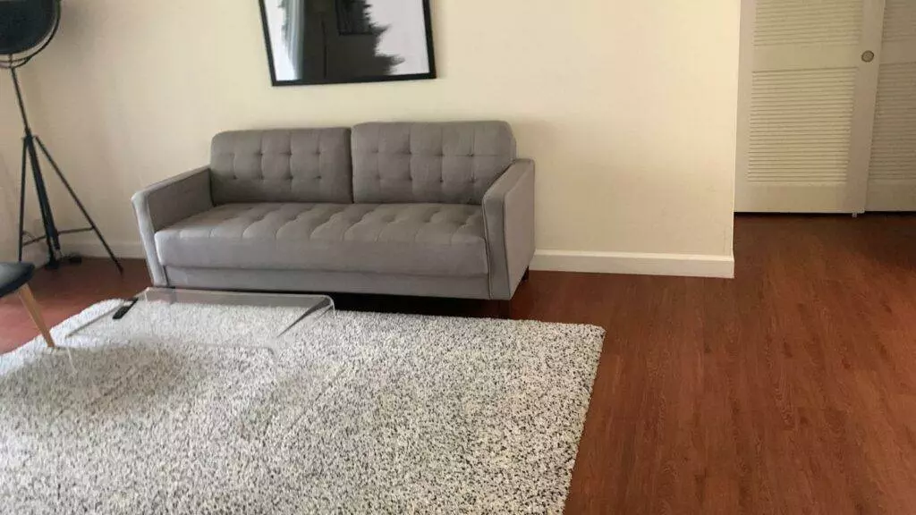 A modern living room featuring a gray tufted sofa, speckled area rug, and hardwood floor, with minimalistic decor and a tripod lamp, recently revitalized by Newark Deep Cleaning.