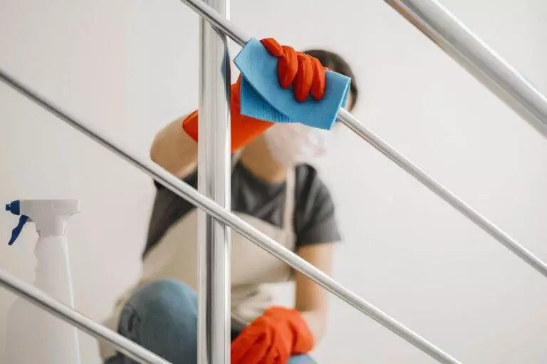 A person wearing red gloves and a face mask cleans a metal handrail with a blue cloth; nearby, a spray bottle sits ready. Master Clean offers customized cleaning services for Los Gatos residents to ensure every detail sparkles.