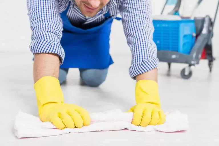 A person in yellow gloves and an apron is kneeling on the floor, diligently cleaning it with a cloth. A Master Clean cart is visible in the background.
