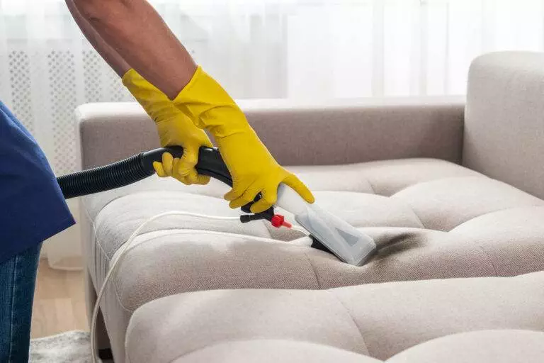 A person wearing yellow gloves uses a steam cleaner to perform upholstery cleaning on a beige upholstered couch.