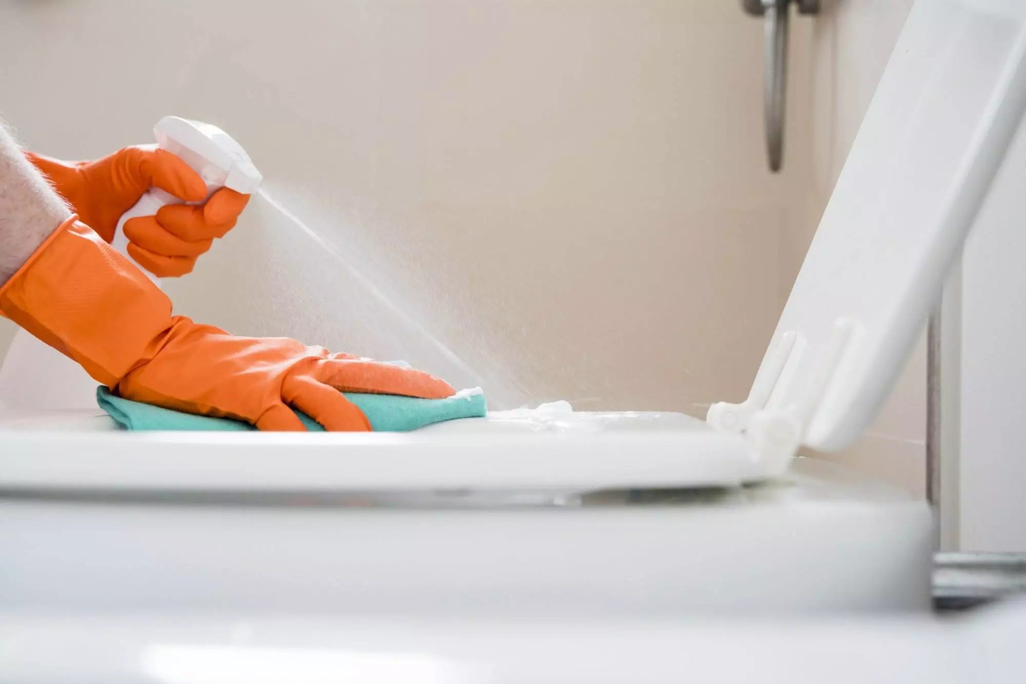 A person wearing orange gloves sprays Bathroom Sparkling Clean fluid onto a toilet seat and wipes it with a green cloth.