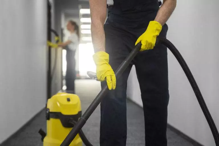 A person wearing yellow gloves operates a vacuum cleaner in a hallway, ensuring a spotless home, while another individual in the background cleans a wall.