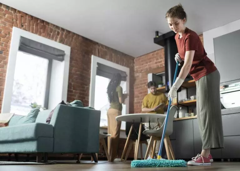A woman mops the floor in a modern living room with exposed brick walls, while a man and another woman, likely Cupertino residents preparing for move-out cleaning, stand in the background near a table.