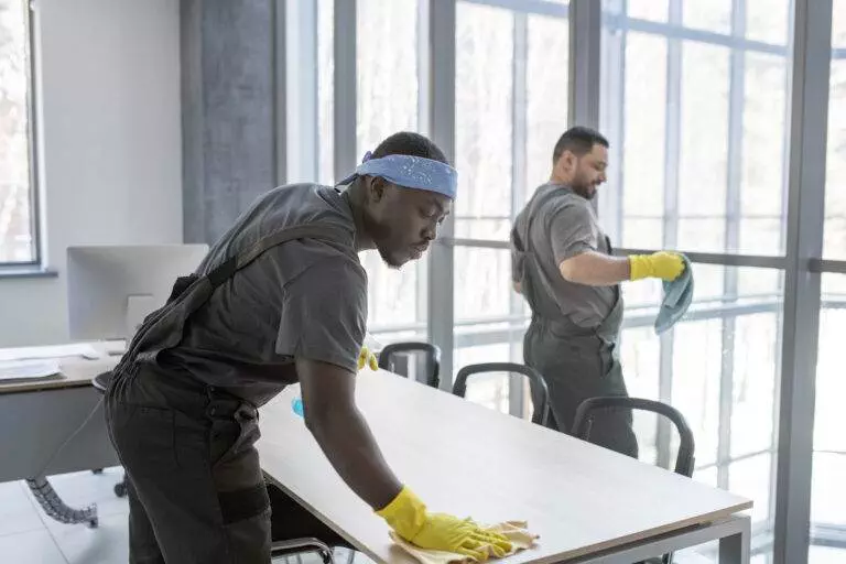 Two janitors from Master Clean, wearing gloves and work uniforms, are meticulously cleaning a table and a window in a bright Saratoga office space. One uses a cloth on the window while the other wipes down the table.