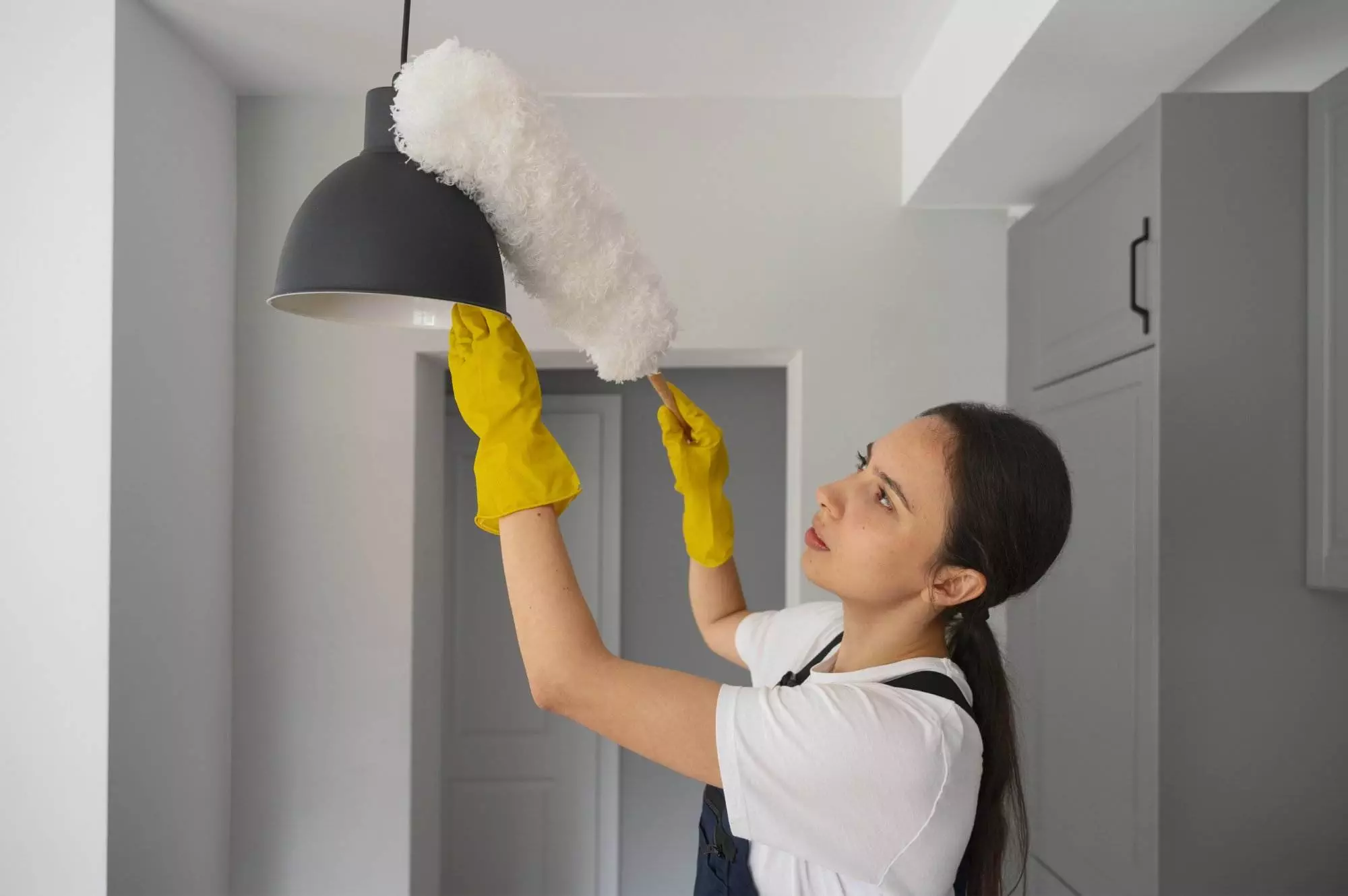 A person wearing yellow gloves and a white shirt cleans a black hanging light fixture in a San Carlos kitchen with a dusting tool, showcasing the meticulous attention to detail provided by Master Clean’s professional room cleaning services.