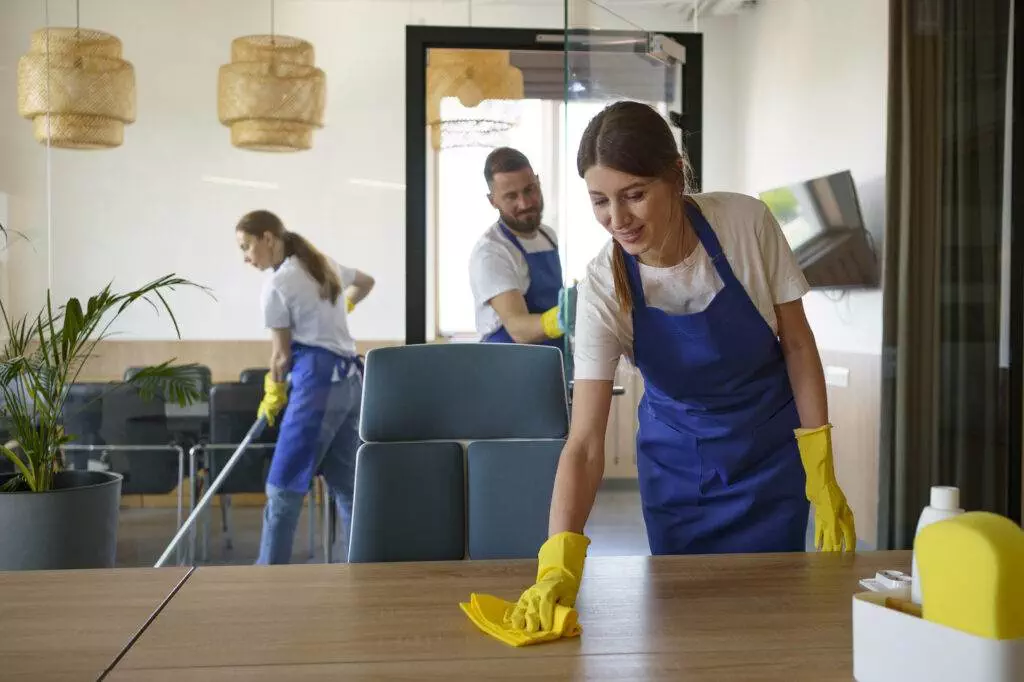 Three people from Master Clean, clad in blue aprons and yellow gloves, are efficiently cleaning an office space in Mountain View; one is wiping a table, another is vacuuming, and the third is cleaning a glass window. For such dedicated attention to detail, consider Full Home Cleaning Services by Master Clean.