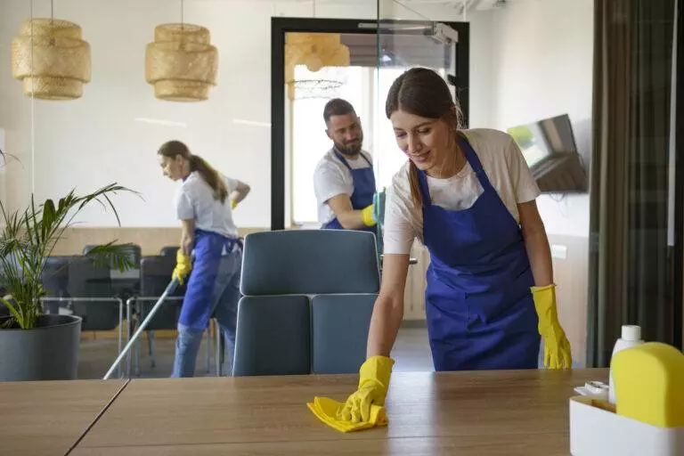 Three people from Master Clean, clad in blue aprons and yellow gloves, are efficiently cleaning an office space in Mountain View; one is wiping a table, another is vacuuming, and the third is cleaning a glass window. For such dedicated attention to detail, consider Full Home Cleaning Services by Master Clean.