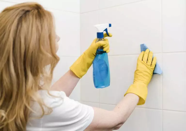 A person wearing yellow gloves is executing professional bathroom cleaning by meticulously scrubbing a tiled wall with a blue spray bottle and a blue cloth, ensuring Belmont Homes maintains its high standards.