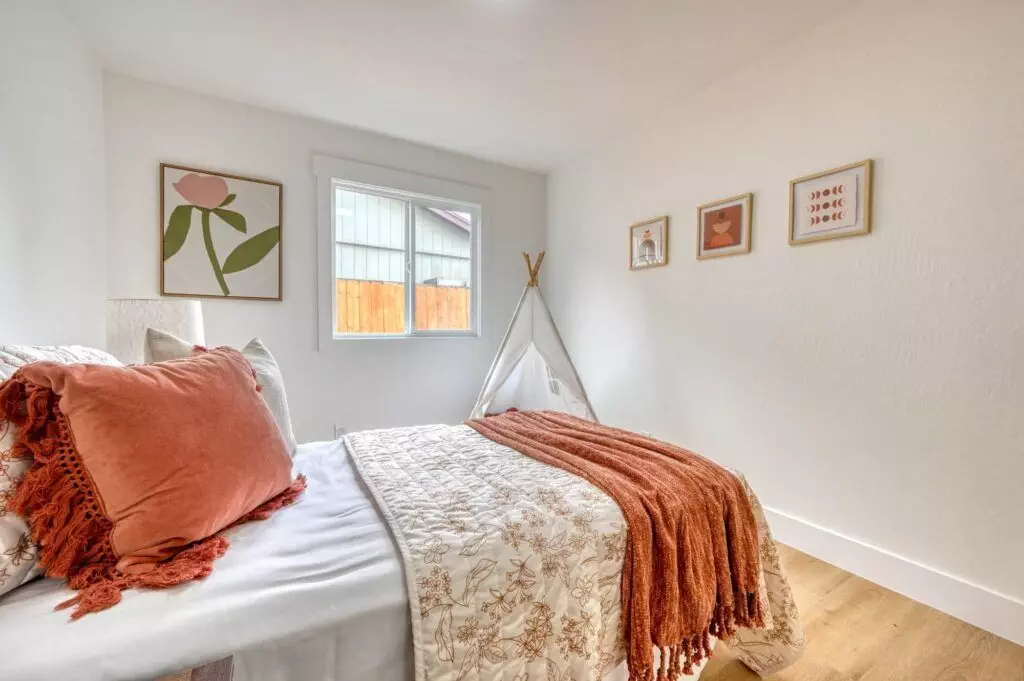 A small, neatly arranged bedroom with a bed, rust-colored throw blanket, decorative pillows, a window, wall art, and a children's teepee in the corner.