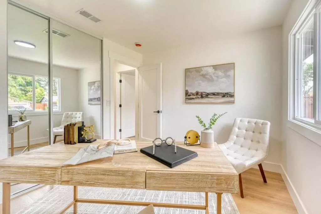 A home office with a wooden desk, a chair, books, decorative items, and artwork on the wall. A large window and mirrored closet doors reflect natural light, illuminating the space.