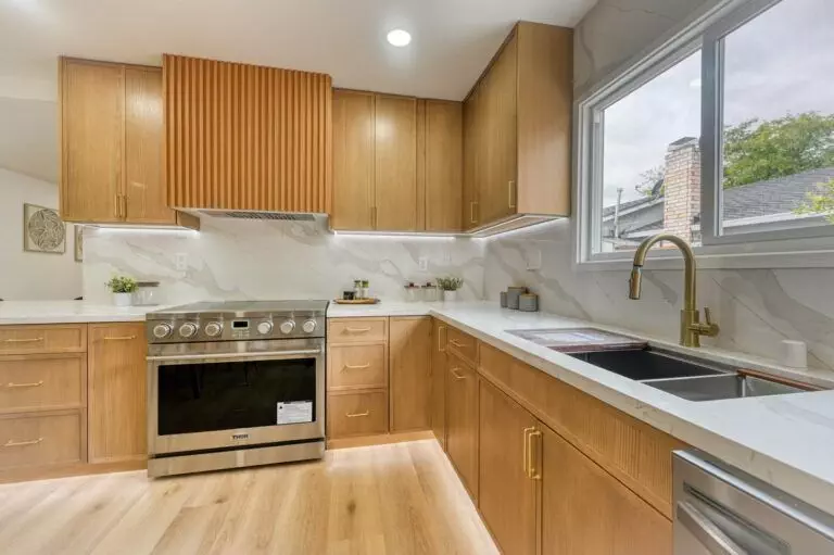 Modern kitchen with wooden cabinets, marble countertops, stainless steel appliances, a large window, and a dual sink with a brass faucet.