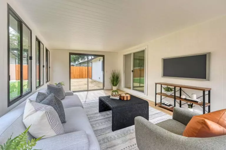 Bright sunroom with large windows, a light grey L-shaped sofa, a black coffee table, and a flat-screen TV on a stand. The room overlooks a fenced backyard with sliding glass doors.