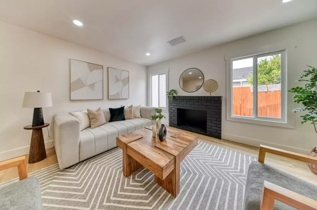 A modern living room with a beige sofa, two armchairs, a wooden coffee table, a fireplace, and a large window. Abstract art and a round mirror decorate the walls. A geometric patterned rug covers the floor.