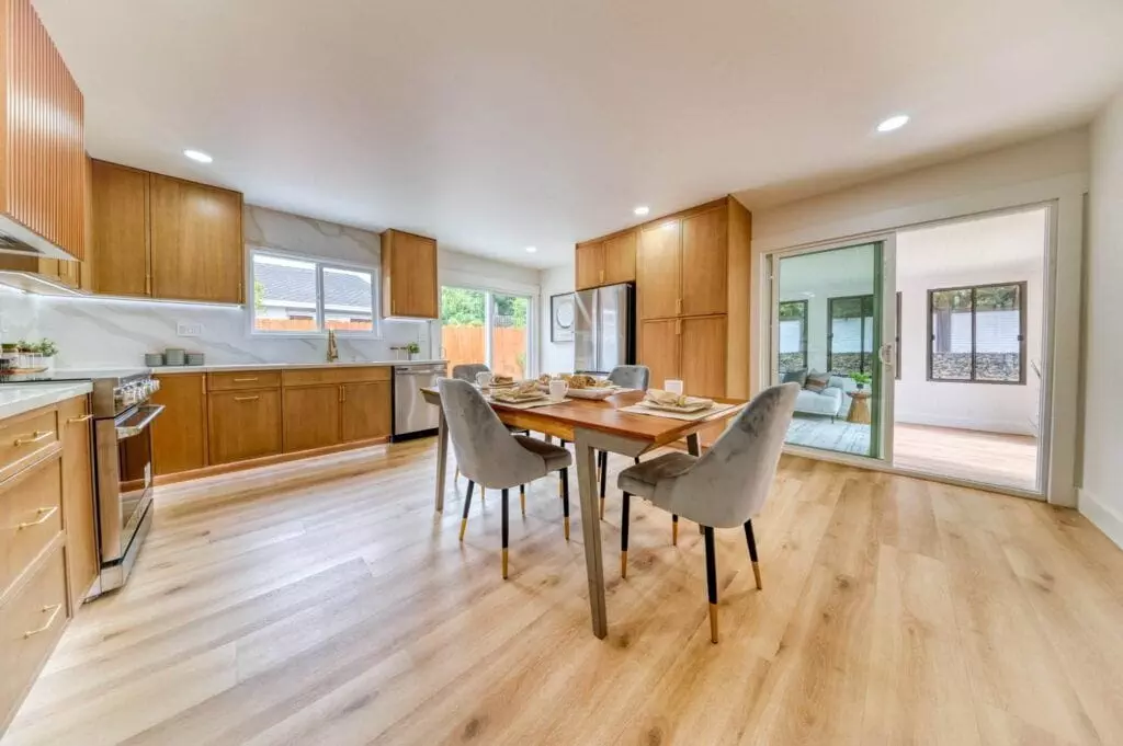 A modern kitchen with wooden cabinets, a dining table set for four, stainless steel appliances, and light wood flooring, adjacent to a sunroom with sliding glass doors.