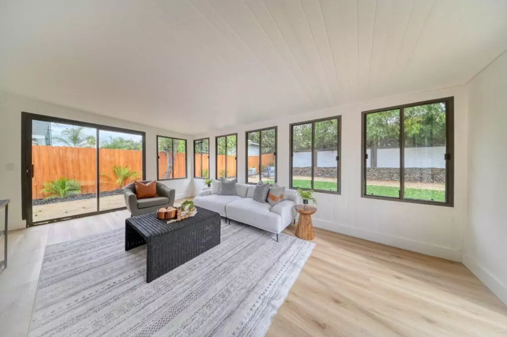 Bright sunroom featuring a comfortable seating area with a white sofa, a gray armchair, and a wicker coffee table. Large windows and glass doors offer views of the fenced backyard and garden.