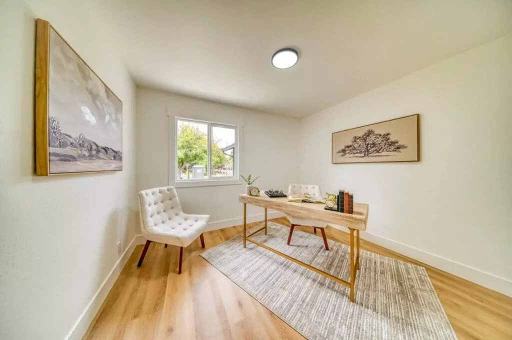 A minimalist home office features a wooden desk, a white cushioned chair, a rug, light-colored walls, two framed artworks, and a window allowing natural light into the room.