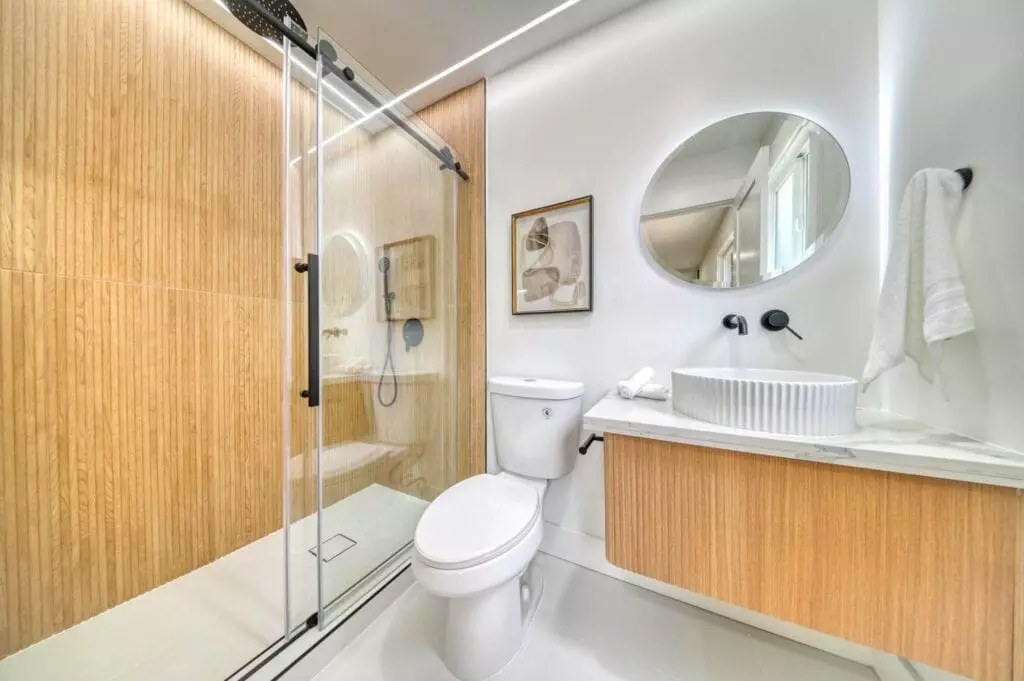 A modern bathroom with a wooden-paneled shower, glass door, white toilet, circular mirror, wall-mounted sink with a wooden base, and white towels.