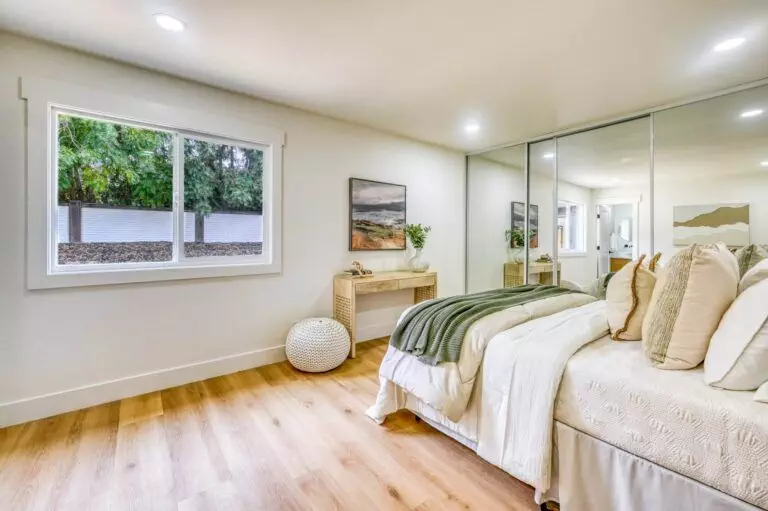 A bright bedroom features a bed with neutral-toned bedding, a window overlooking greenery, a mirrored closet, a small wooden desk, and a round woven basket on a wooden floor.