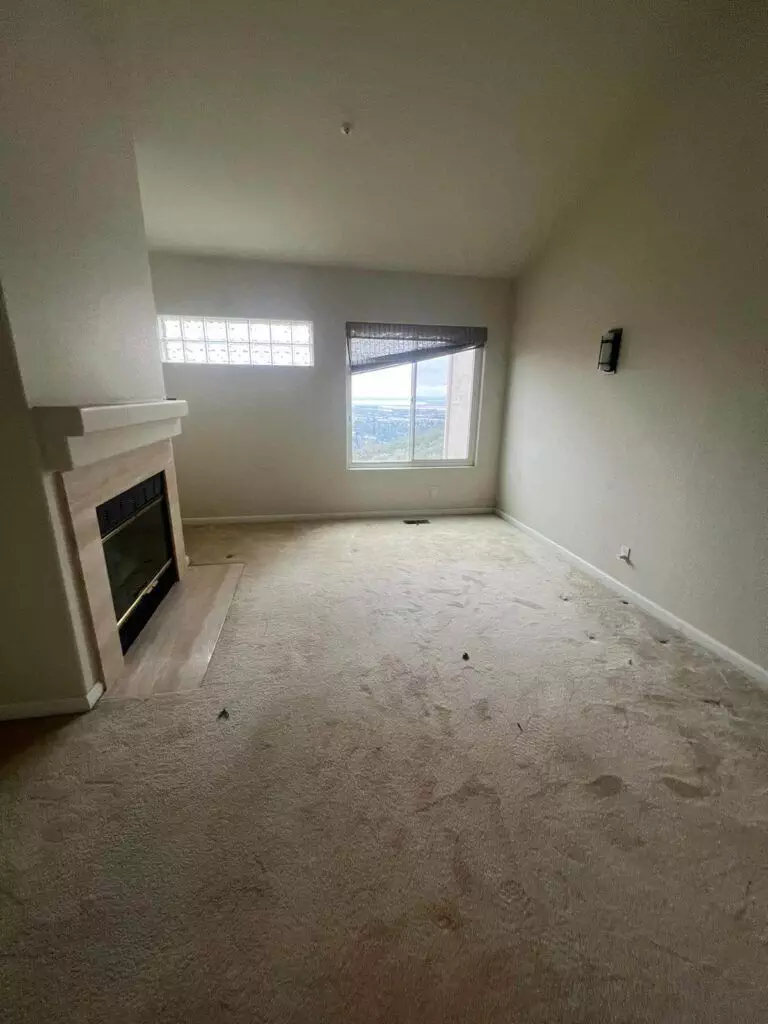 A sparsely furnished room with beige carpet, a fireplace on the left, a small window above it, and a larger window with a view on the right wall. The room has high ceilings and appears empty.