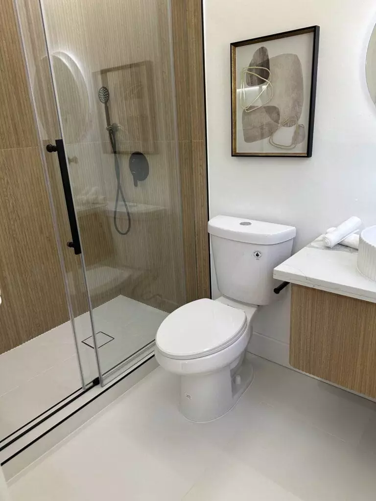 A modern bathroom with a glass-enclosed shower, a toilet, a framed abstract artwork, and a vanity with a round mirror and white sink.